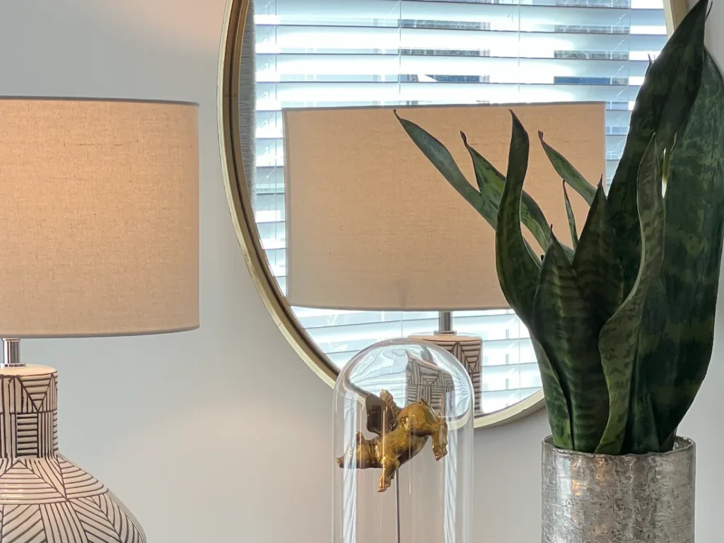 A homeowner has opted for home staging services and the designer implented a modern, earth-toned lamp shade with the base featuring geometric patters. Also pictured is a snake plant and a decorative golden pig with wings on a stand inside a glass dome.